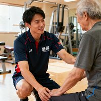 一位三十岁出头的男性社会体育指导员穿着新球体育制服，在训练区单膝跪地为一位老年居民调整运动姿势，表情专注耐心